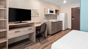Habitación de hotel compacta con cama, escritorio con televisor de pantalla plana, silla, kitchenette con microondas y refrigerador, y una puerta.