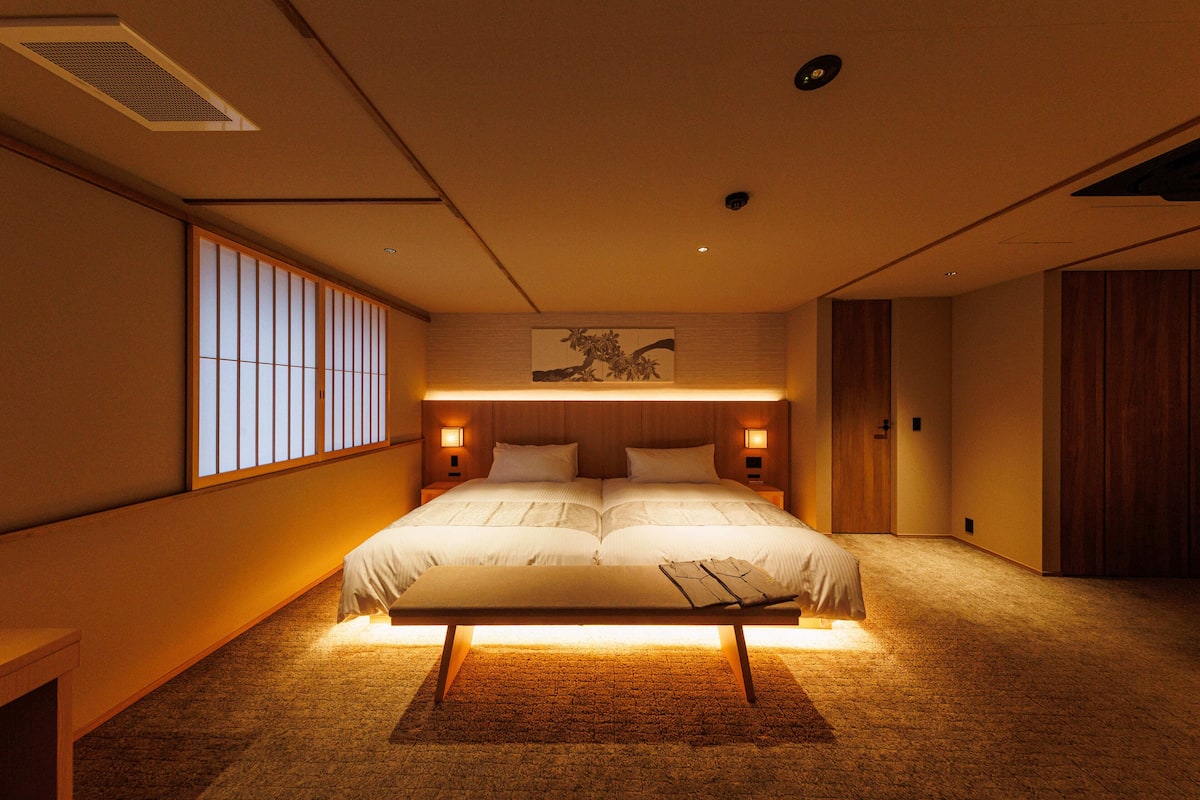 A modern bedroom with a large bed, bedside lamps, a wooden headboard, and a view of the outside through a sliding door.