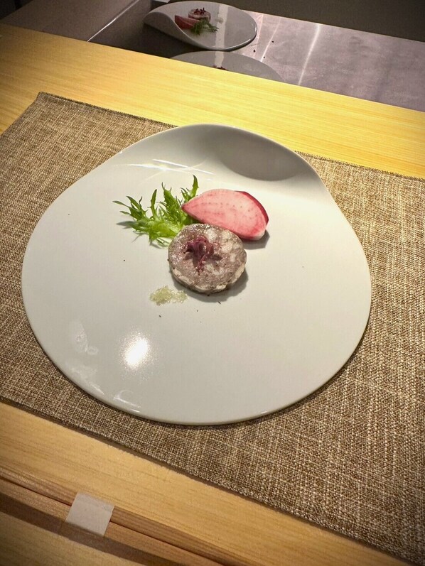 Une assiette avec une assiette blanche ronde, un morceau de viande, un radis et quelques feuilles de salade, le tout posé sur une table recouverte d’un set de table en fibres tressées.