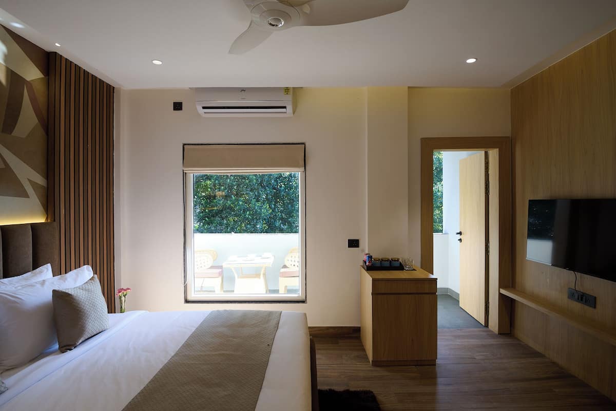 A modern hotel room with a large bed, a small desk, a TV, and a balcony with a view of greenery.
