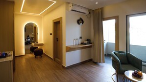 Et moderne hotelværelse med trægulv, et siddeområde med en grøn lænestol, et lille bord med frugt, et spejl og en køkkenbordplade med kaffemaskine.