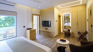 Et moderne hotelværelse med en stor seng, et fjernsyn ophængt på væggen, et lille bord med frugt og et siddeområde med en stol og skammel.