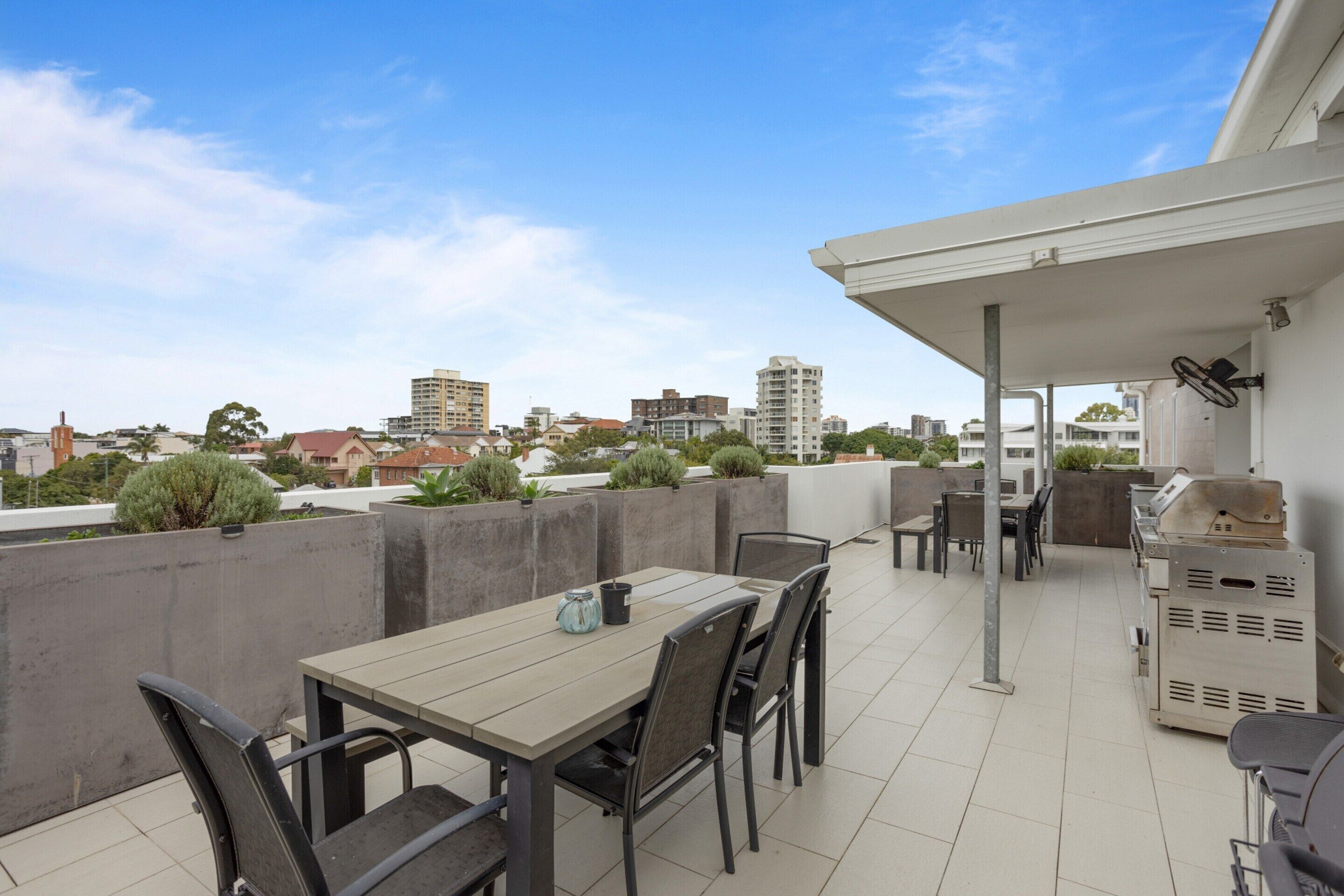 A rooftop patio with a table and chairs, a grill, and a view of the cityscape.