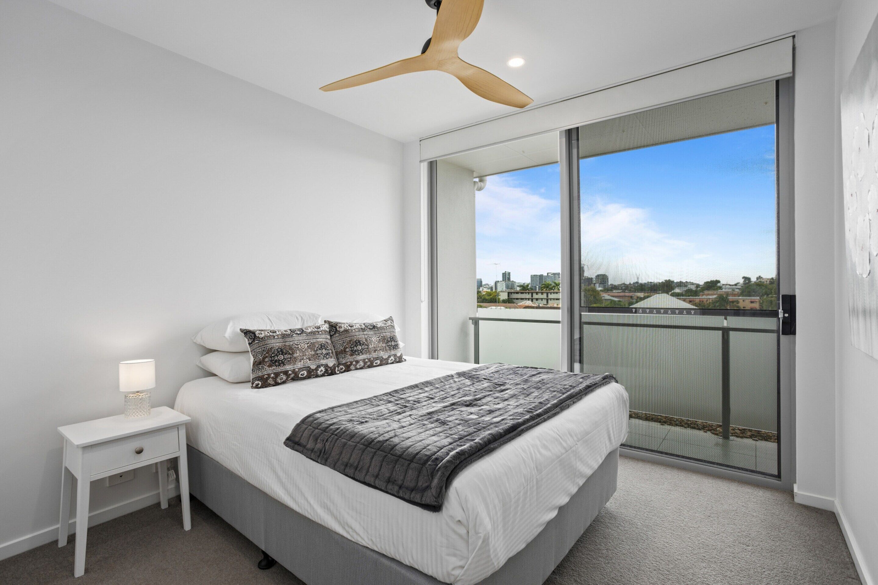 A bedroom with a bed, nightstand, and ceiling fan. It has a large window with a view of buildings and a balcony.