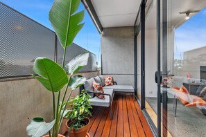 Ein moderner Außenbereich mit Holzterrasse, einer grauen Couch und orange-weißen Kissen sowie großen, grünen Pflanzen.