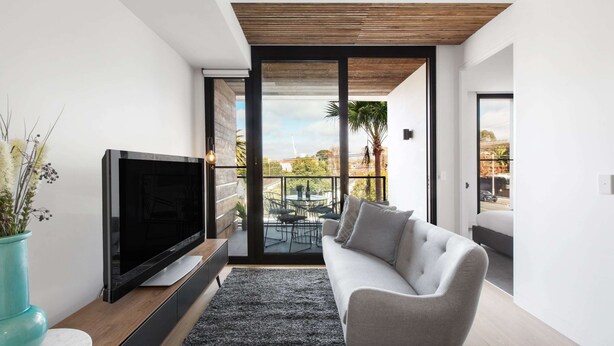 A modern living room with a flat-screen TV, a grey sofa, and a large window overlooking a balcony with outdoor furniture. - Chic 1-bed With a Sleek Balcony Near Shops (Melbourne)
