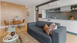 A modern living room with a grey sofa, a dining area with a white table and brown chairs, and a kitchen with white cabinets and blue tiles. - Modern 1-bed, City Skyline Views & Close to Shops (Melbourne)