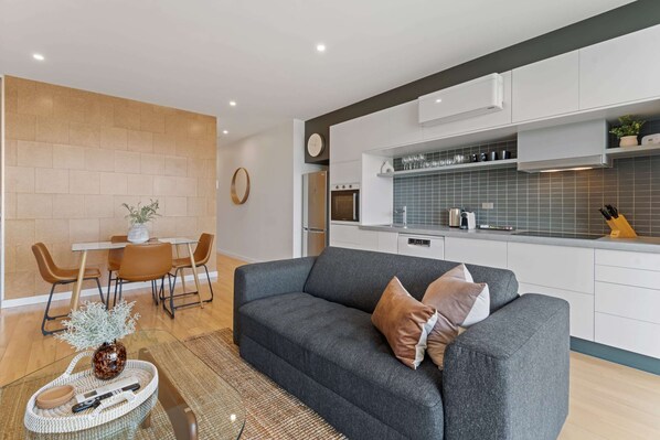A modern living room with a grey sofa, a dining area with a white table and brown chairs, and a kitchen with white cabinets and blue tiles. - Modern 1-bed, City Skyline Views & Close to Shops (Melbourne)