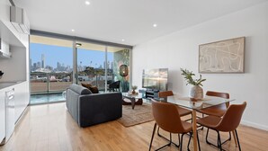 A modern living room with a glass dining table, grey sofa, and a city view through large windows. - Modern 1-bed, City Skyline Views & Close to Shops (Melbourne)