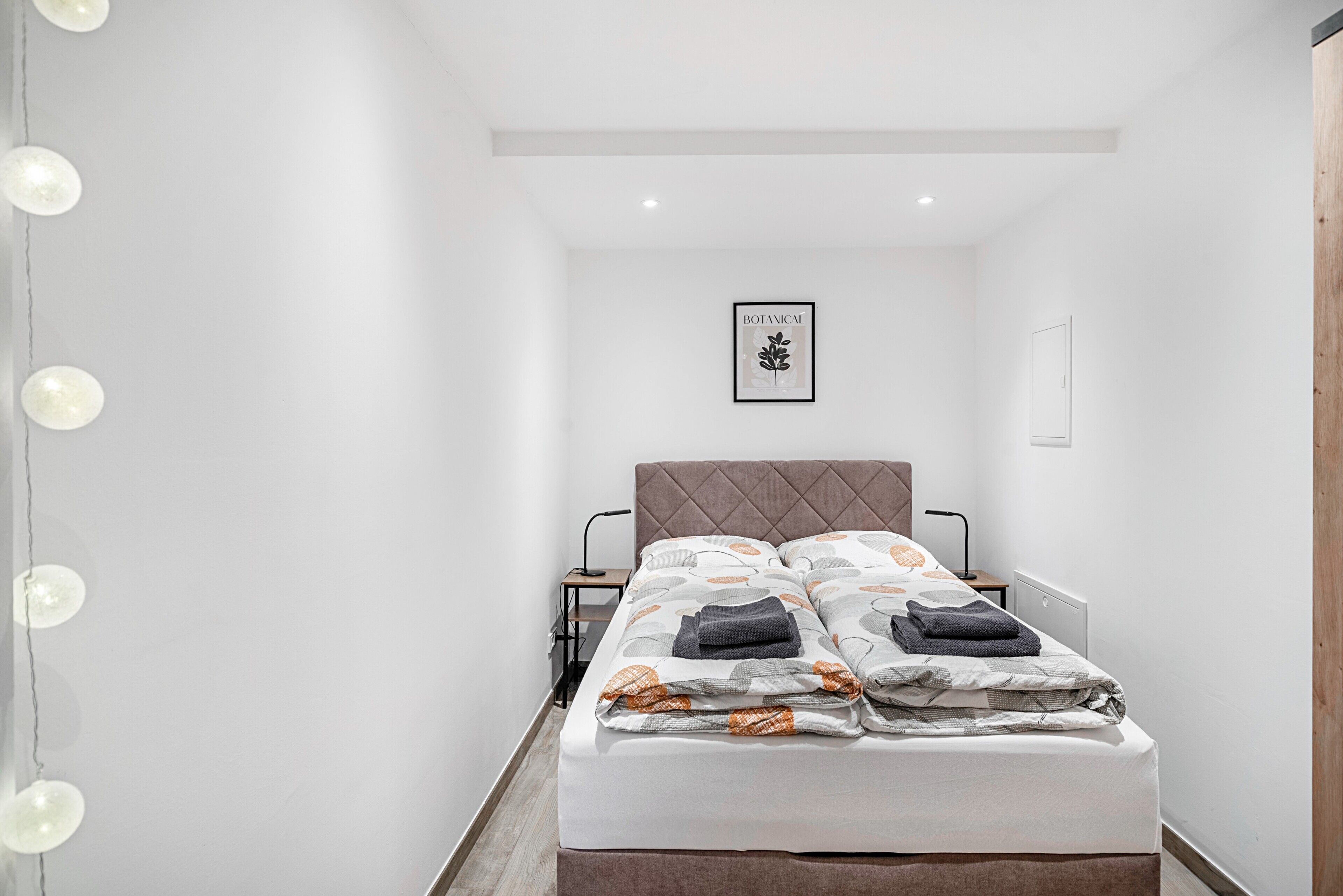A neatly made bed with a pillow and a blanket, a bedside table with a lamp, and a framed botanical print on the wall.