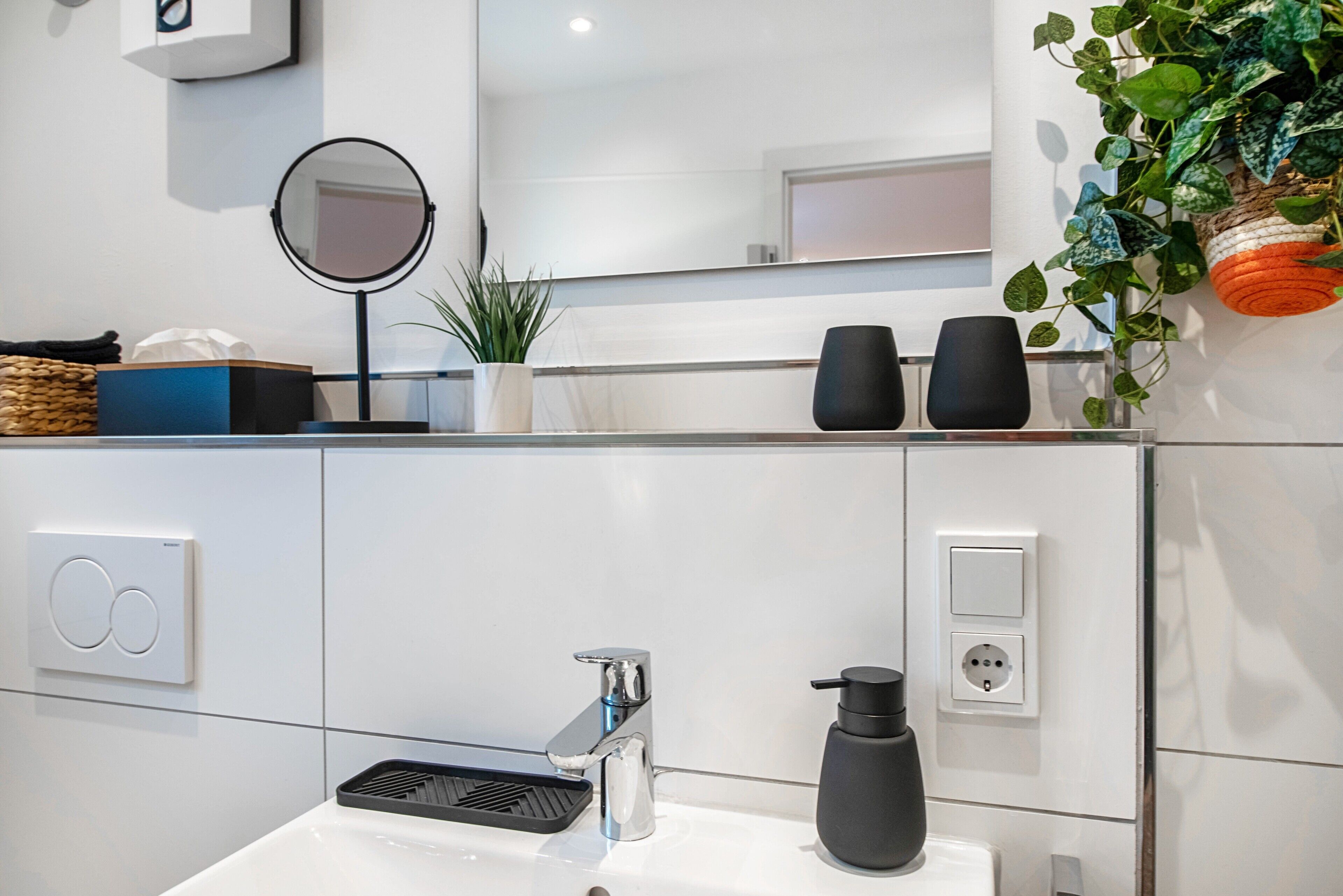 Een moderne badkamer met een witte wasbak, een spiegel, een zwarte zeepdispenser en een plant in een pot.