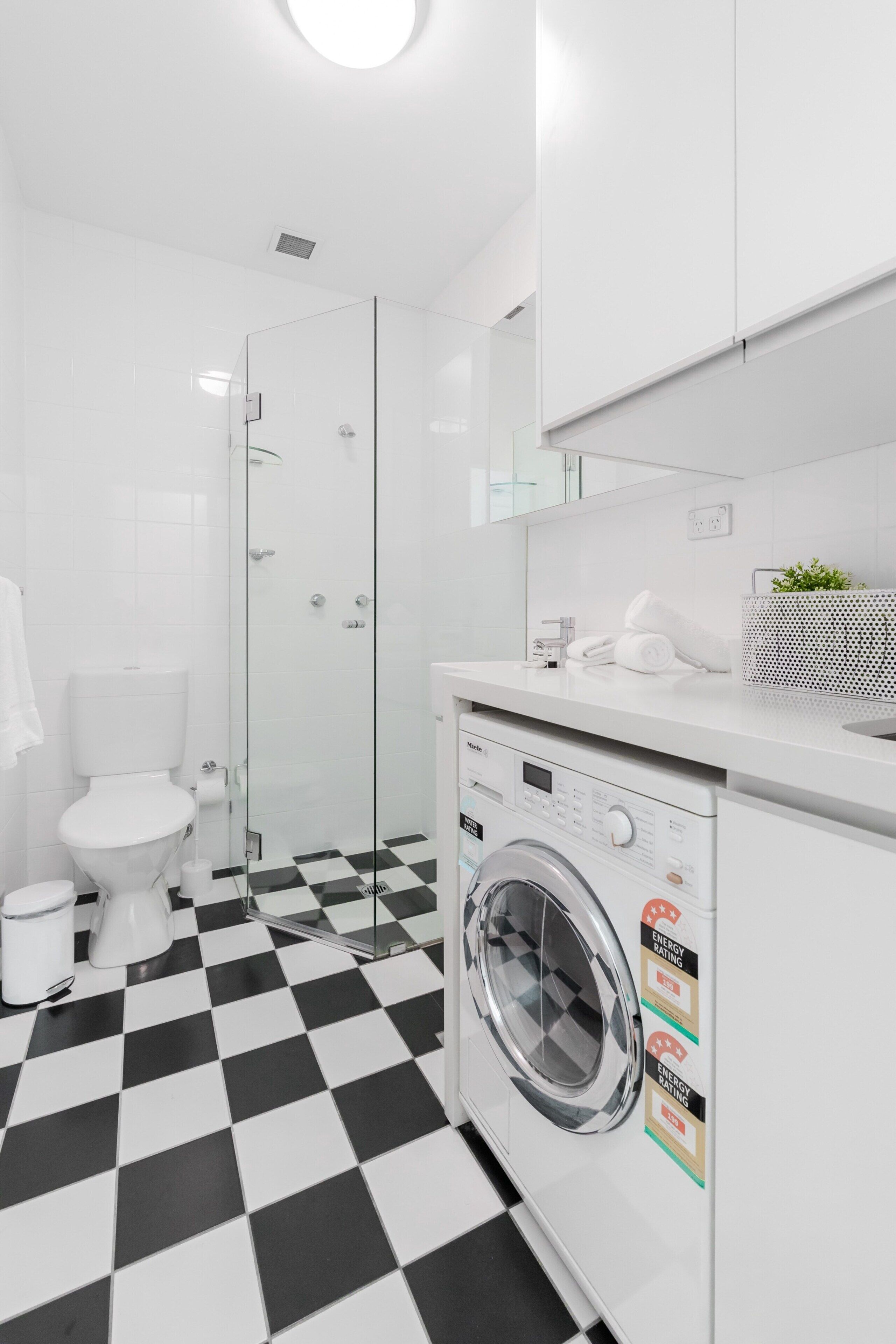 Ein modernes Badezimmer mit schwarz-weiß kariertem Boden, einer Duschkabine aus Glas, einer weißen Toilette und einer Frontlader-Waschmaschine.