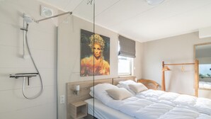 A modern bedroom with a glass shower enclosure, a large bed with white bedding, a wooden nightstand, and a wall-mounted artwork.