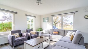 A spacious living room with a grey sofa, armchairs, and a coffee table. Large windows provide natural light and views of the outdoors.