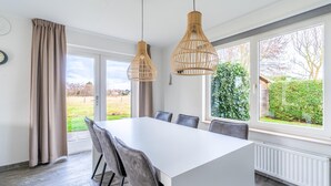 A modern dining area with a large white table, grey upholstered chairs, and two wicker pendant lights.