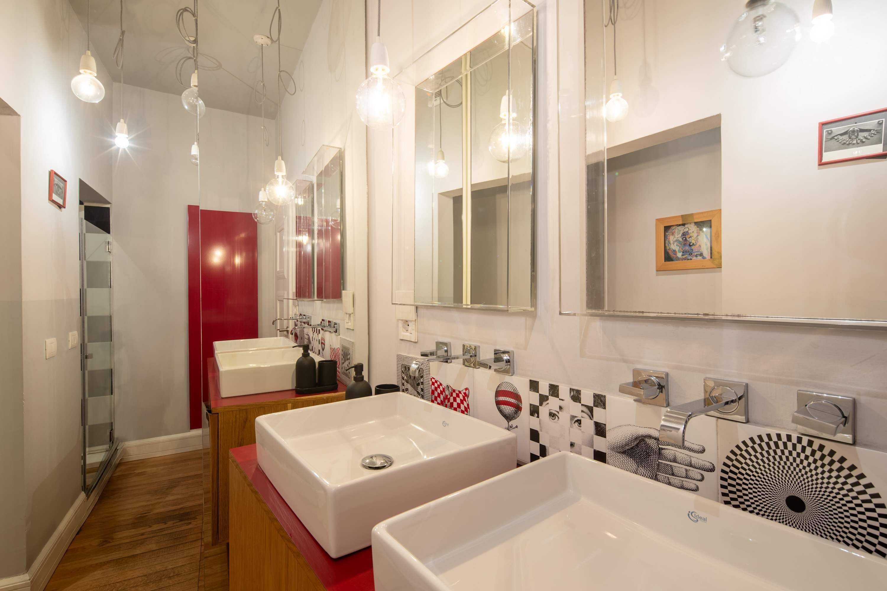 A modern bathroom with two large white sinks, a red accent wall, and a mirror with a checkered pattern.