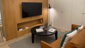 Egy modern nappali, melyben egy síkképernyős televízió van a falra szerelve, a fal pedig fából készült. A szobában található még egy fekete kávézóasztal, rajta könyvekkel és egy vázzával, egy barna kanapé és egy szekrény.