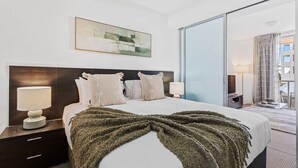 Una habitación de hotel moderna con una cama grande, dos lámparas de noche, un cabecero oscuro, un cuadro sobre la cama y una puerta corrediza de vidrio que da a un balcón.