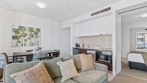 Una sala moderna con un sofá gris, una zona de comedor con mesa y sillas, una cocina con armarios blancos y un dormitorio con cama y ventana.