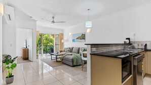 A modern kitchen with a central island, a dining area with a sofa and table, and a view of greenery through the glass door. - Urban Tropics Modern Resort Apartment (Cairns)