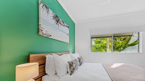 A bedroom with a bed, white bedding, a nightstand with a lamp, a decorative shell, and a painting of a beach scene. - Urban Tropics Modern Resort Apartment (Cairns)
