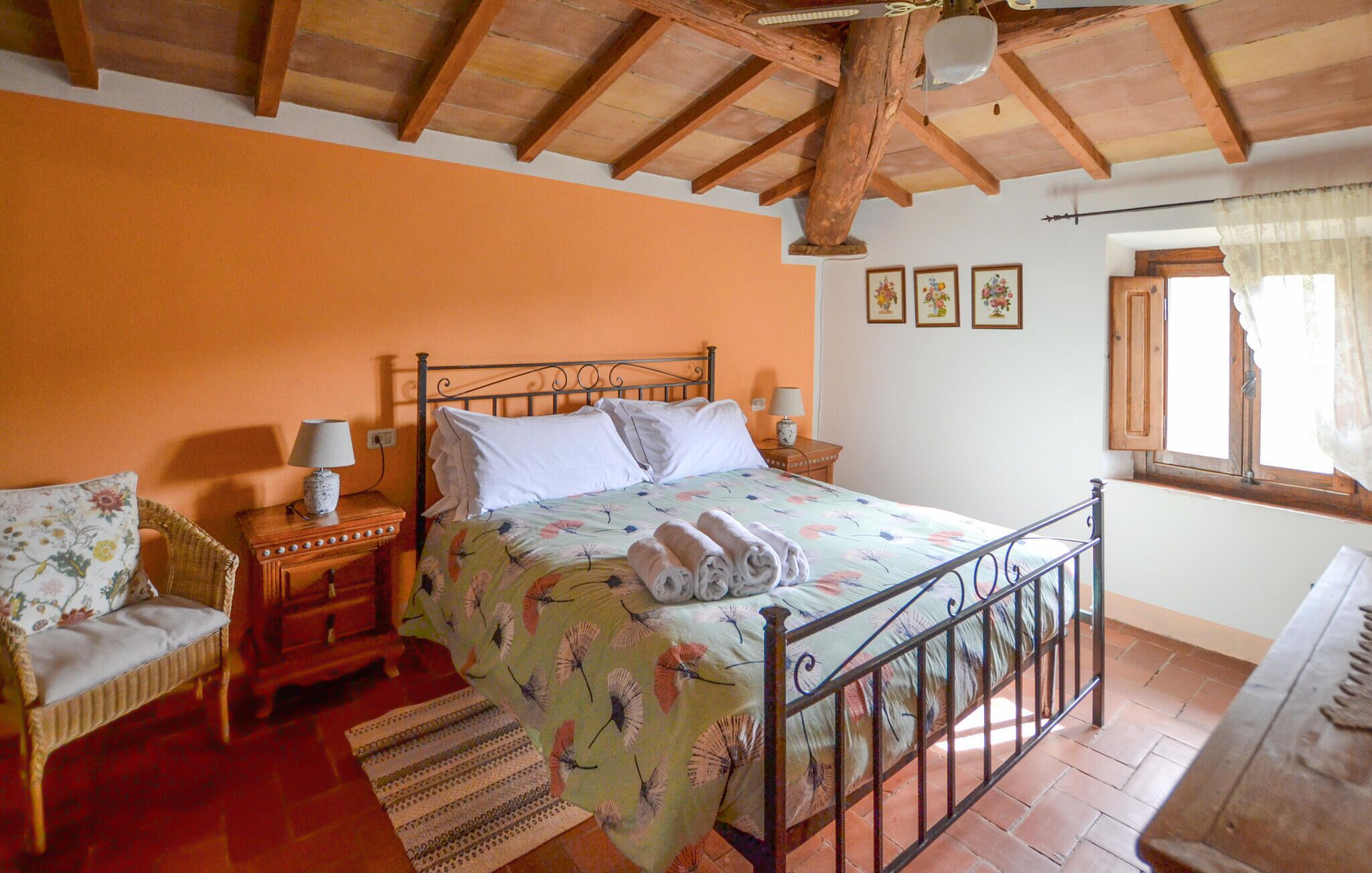 A bedroom with a wooden ceiling, a bed with a floral bedspread, two bedside tables with lamps, a wicker chair, and a window with curtains.