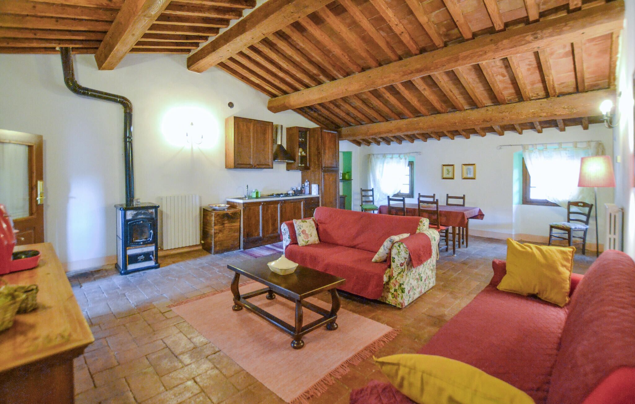A cozy living room with a red sofa, a wooden coffee table, and a fireplace. There is a kitchen area with wooden cabinets and a dining table.