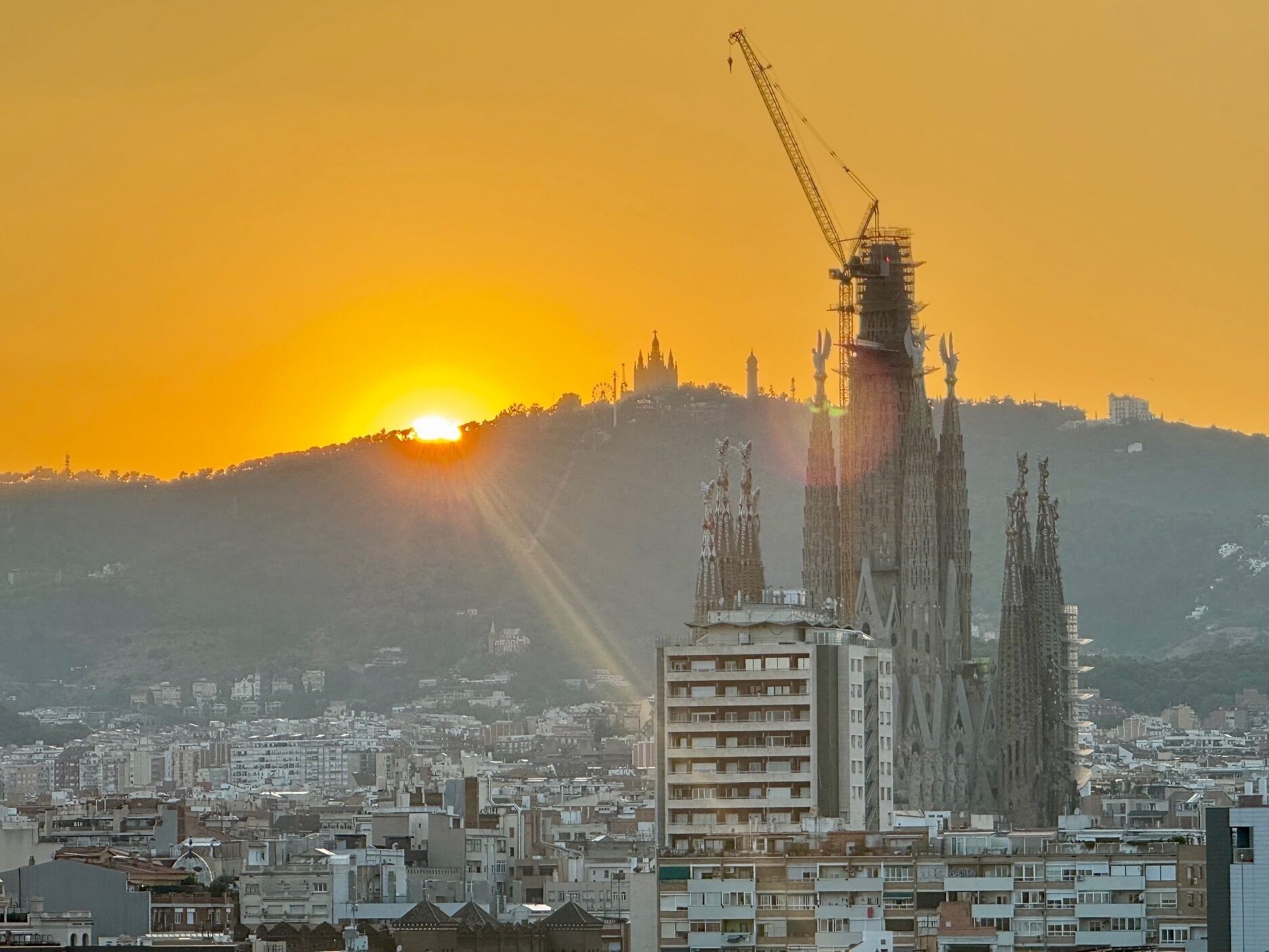 Un paisaje urbano con un edificio en construcción destacándose, mostrando varias grúas y una vista despejada del sol poniéndose al fondo.