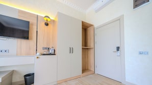 A modern room with a flat-screen TV mounted on the wall, a wooden cabinet with a built-in shelf, and a white wardrobe with a door. - Ayçiçeği Comfort Hotel (Edirne)