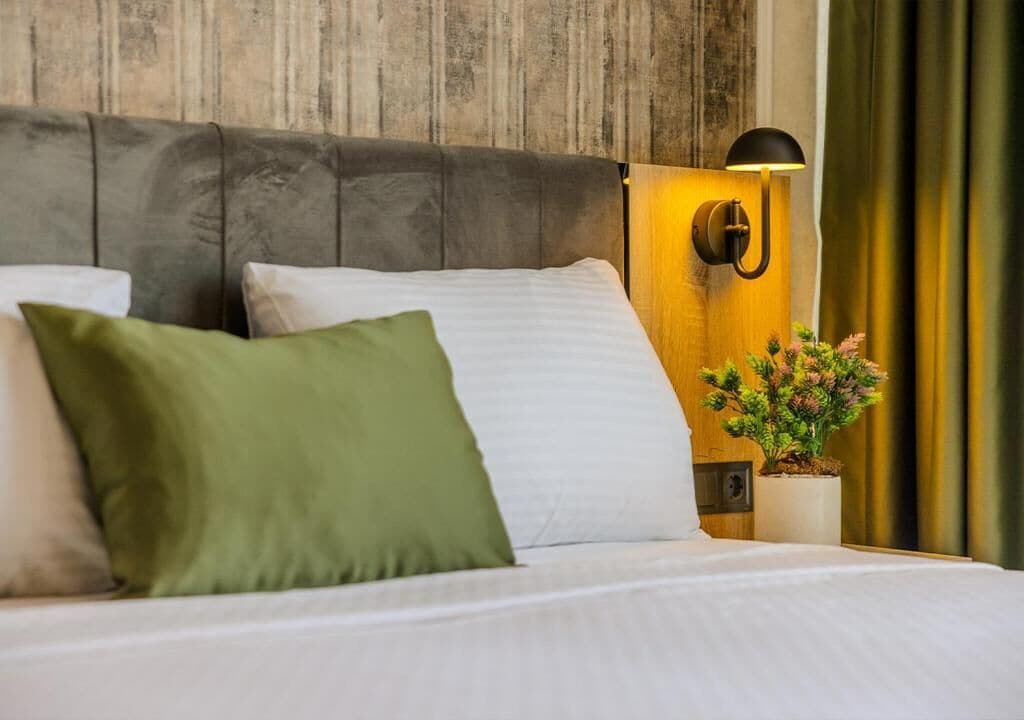 A neatly made bed with white and green pillows, a wooden headboard, a wall-mounted lamp, and a small potted plant.