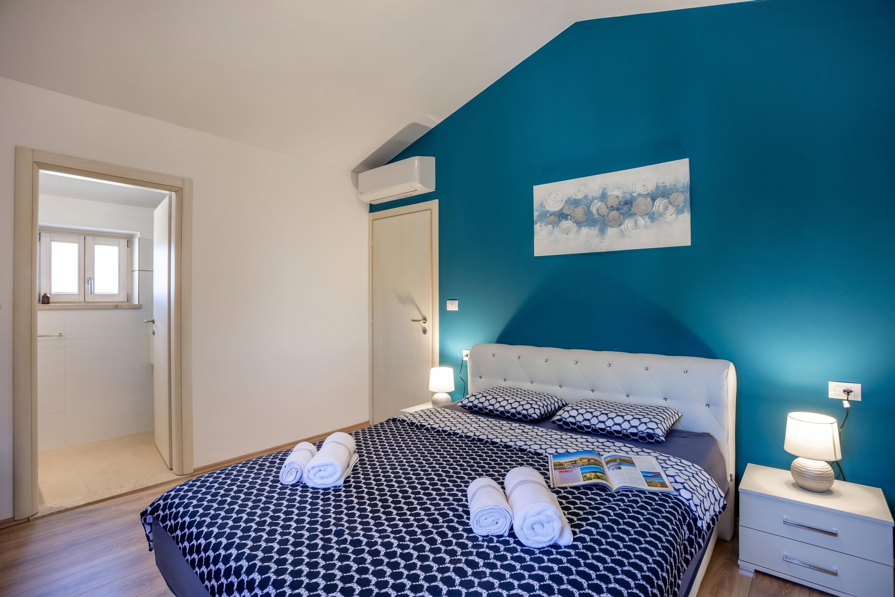 A hotel room with a bed, bedside tables, a nightstand, a wall-mounted air conditioner, and a door leading to another room.