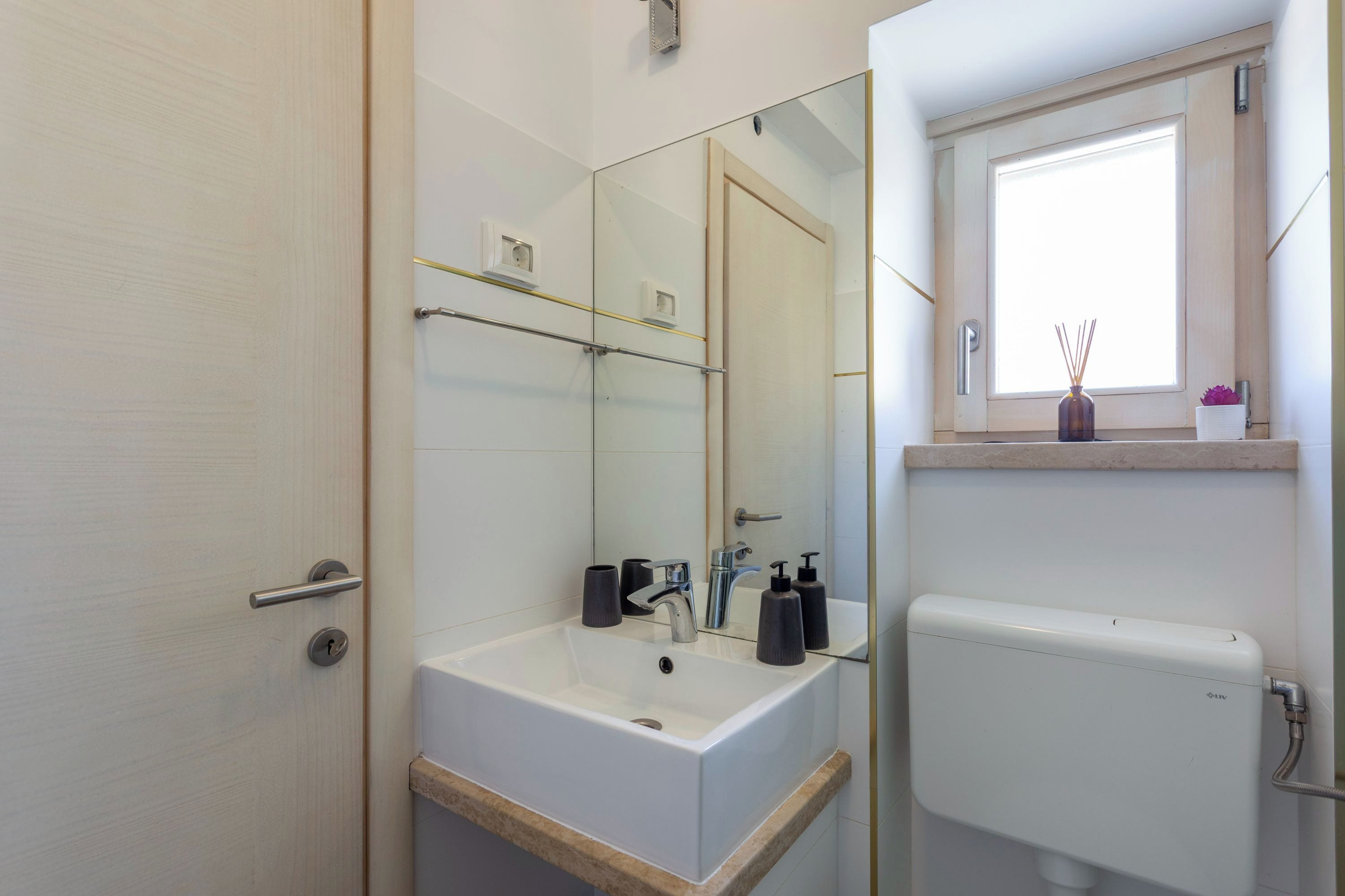 Ein modernes Badezimmer mit einem weißen Waschbecken, Spiegel und Toilette. Über der Toilette befindet sich ein Fenster mit Ablage.