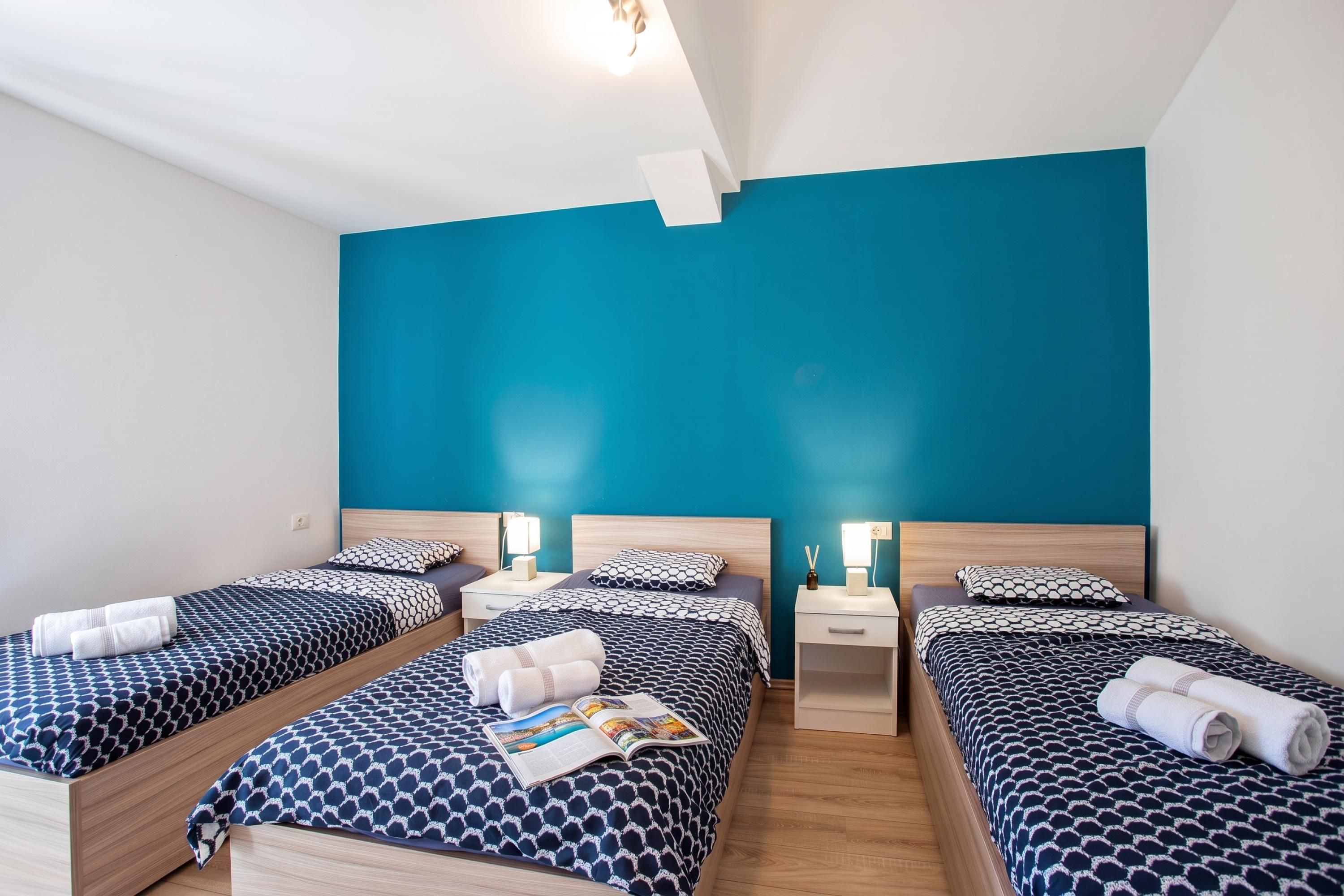 A hotel room with three beds, each with a patterned bedspread, a nightstand with a lamp, and a wall with a solid color.
