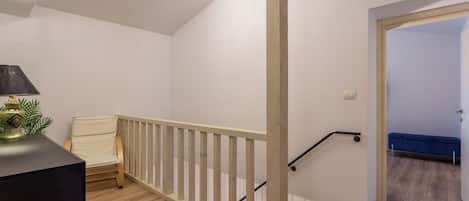 A staircase with a wooden handrail and a black handrail, leading to a room with a blue bench and a white chair.