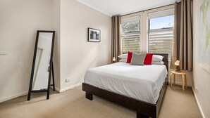 A bedroom with a bed, a mirror, a small table with a lamp, and a window with curtains. - Stunning 2-bed Unit With BBQ Patio Near Dining (Melbourne)