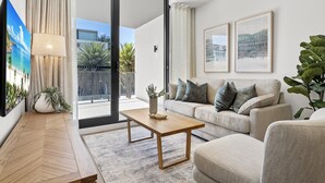 A modern living room with a sofa, a coffee table, and a TV. There is a sliding glass door leading to a balcony with plants. - Fremantle 1-bed Close to Beach & Shops (Perth)