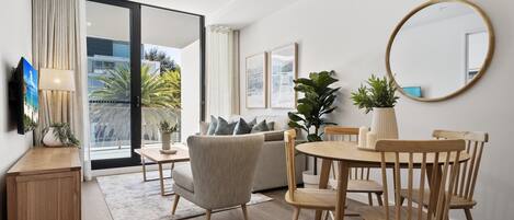 A modern living room with a sofa, dining table, and chairs, a TV, and a large window with a view of palm trees.