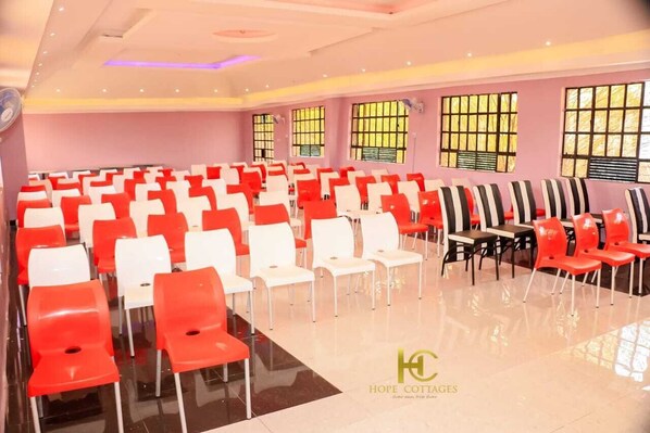 A conference room with rows of chairs, large windows, and a logo on the wall. - Hope Cottages and Recreation Centre (Marigat)