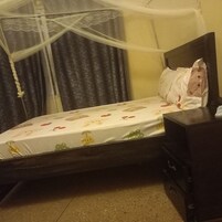 Una cama con colcha floral, cabecera de madera, mesita de noche con cajones y mosquitera sobre la ventana.