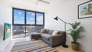 Een moderne woonkamer met een grijze hoekbank, een groot raam met uitzicht op de stad, een kleurrijk gestreept tapijt, een vloerlamp en een potplant.