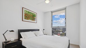 Een slaapkamer met een bed, twee nachtkastjes, een bijzettafeltje, een plafondventilator, een raam met uitzicht op de stad, en een gekaderd schilderij aan de muur.