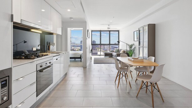 Een moderne keuken met witte kasten, een eethoek met een houten tafel en stoelen, en een uitzicht op de stad door grote ramen.