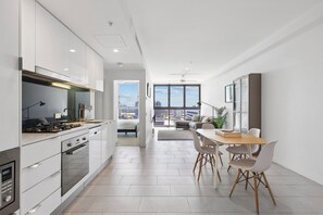 A modern kitchen with white cabinets, a dining area with a wooden table and chairs, and a view of the city through large windows. - (Brisbane)