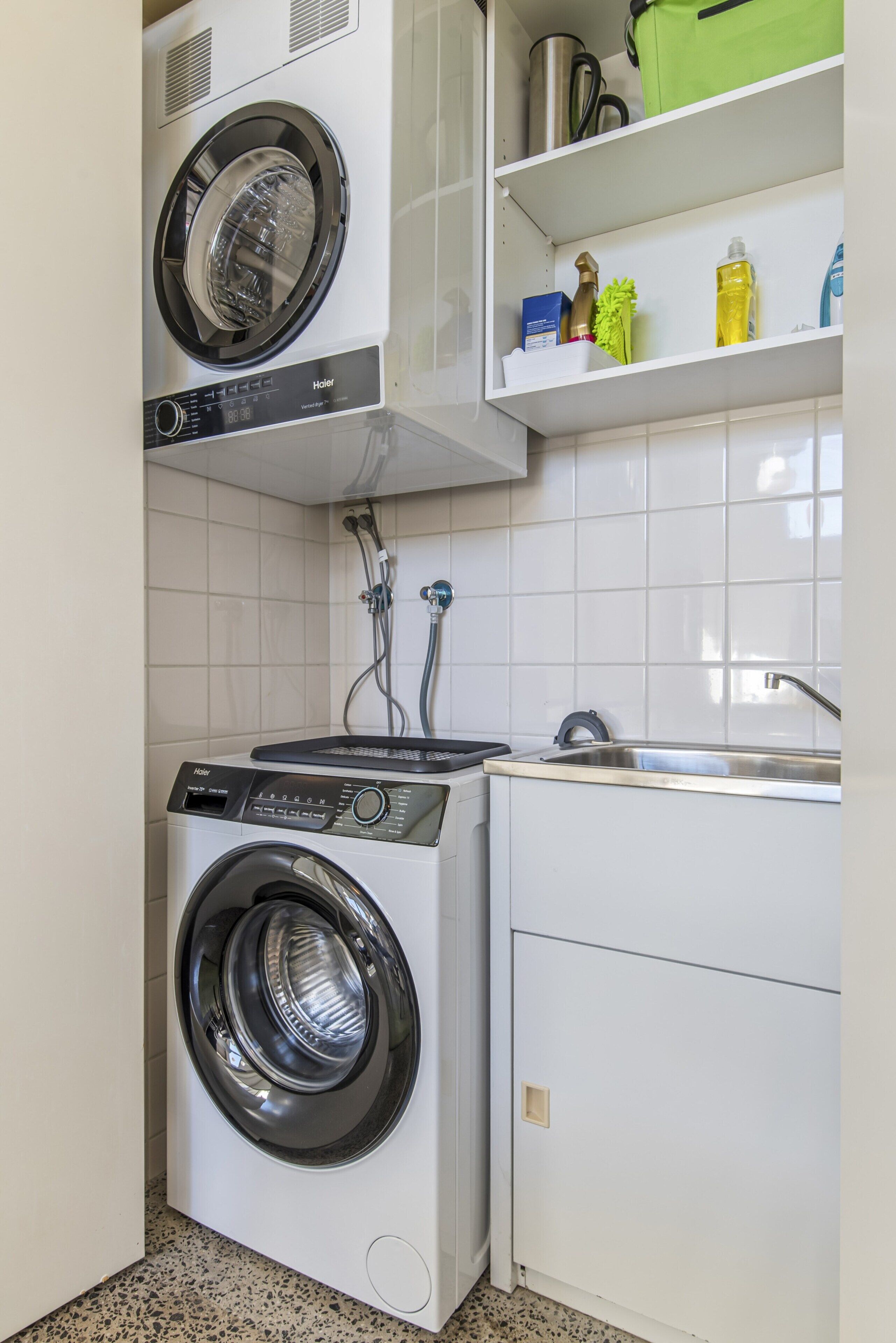 A compact laundry area with a front-loading washer and dryer, a sink, and a shelf with cleaning supplies.