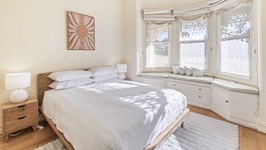Un dormitorio con una cama grande, una mesita de noche, un banco junto a la ventana y una obra de arte en la pared.
