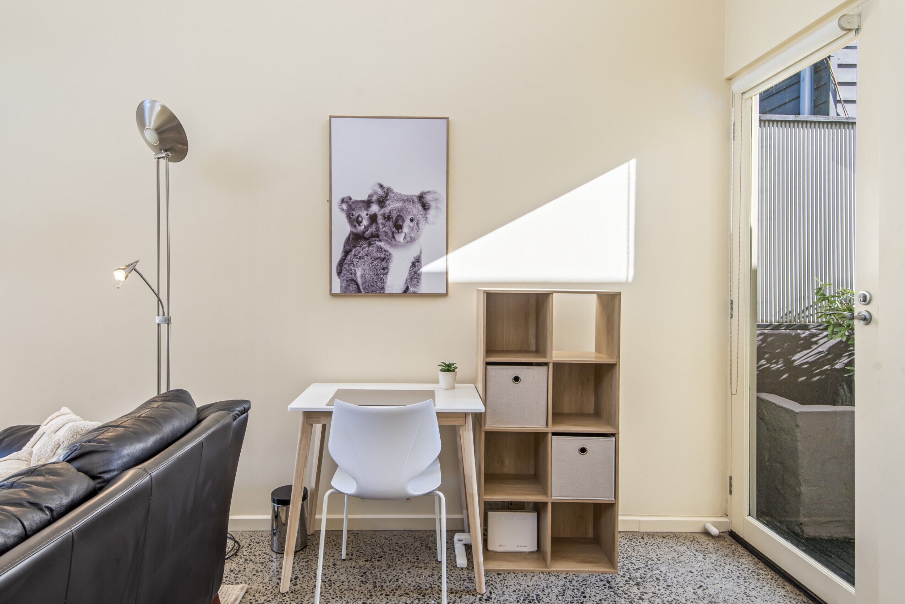 A modern living room with a black leather sofa, a white desk, a white chair, a floor lamp, a framed picture of koalas, and a sliding glass door.