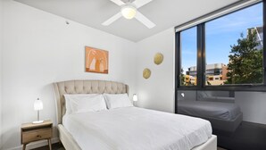 A bedroom with a large bed, a nightstand, a ceiling fan, a window with a view of buildings, and a wall-mounted artwork. - Cosy 2-bed With Pool, Bbq Area and Secure Parking (Brisbane)