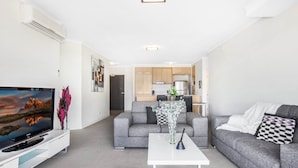 A modern living room with a flat-screen TV, grey sofas, a white coffee table, and a kitchen in the background. - Lakeside Family Oasis With Pool, Gym and Balcony (Gold Coast)