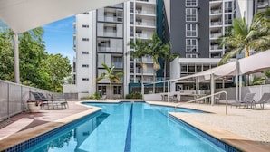 A swimming pool with lounge chairs and umbrellas, surrounded by a modern building with balconies and palm trees. - Lakeside Family Oasis With Pool, Gym and Balcony (Gold Coast)