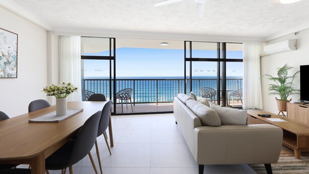A modern living room with a balcony overlooking the sea, featuring a sofa, dining table, and chairs.